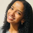 Headshot of a person with curly hair, smiling at the camera, wearing hoop earrings and a grey top.