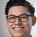 A headshot of a person wearing glasses, smiling, with dark, curly hair, dressed in a collared shirt and jacket.