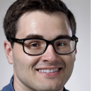 A headshot of a person with short dark hair, wearing black glasses, and smiling. They are dressed in a blue collared shirt.