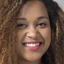 Headshot of a smiling person with curly hair and a nose ring, wearing pink lipstick.