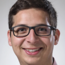 A headshot of a person with short brown hair, wearing black glasses and a pink shirt, smiling at the camera.