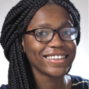 Headshot of a person with braided hair and glasses, smiling at the camera, wearing a patterned top. The background is plain.