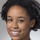 A headshot of a smiling person with natural curly hair, wearing a black top with a subtle pattern, set against a neutral gray background.