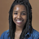 A headshot of a person smiling with long braided hair, wearing a denim shirt over a light-colored top. The background is a plain, warm brown.