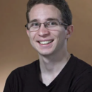 A smiling person wearing glasses and a dark shirt, positioned against a plain, neutral background. Headshot.