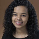 A headshot of a smiling person with curly hair, wearing a black top, set against a brown background.