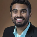 Headshot of a person with short dark hair and a beard, wearing a dark suit jacket and a light blue shirt, smiling against a brown background.