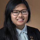 A headshot of a person with long black hair and glasses, wearing a black blazer over a striped shirt, against a plain background.