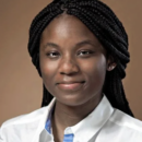 Headshot of a person with braided hair, wearing a white shirt, set against a plain brown background.