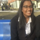 A person with long hair and glasses is sitting on a blue bench, smiling. They are wearing professional attire with a white shirt and dark jacket. In the background, a group of people is gathered near a brick building.