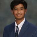 Headshot of a person wearing a blue suit, white shirt, and patterned tie, with a neutral expression against a gray background.