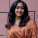 A person with long wavy hair, wearing a bronze blouse, smiling in front of a textured wall. Headshot.