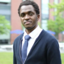 A headshot of a person wearing a navy cardigan, white shirt, and blue tie, standing outdoors with a blurred building and trees in the background.