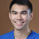 A headshot of a smiling person wearing blue scrubs, set against a plain brown background.