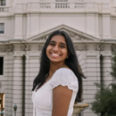 A person smiling in front of an ornate building with classical architecture, wearing a white shirt and standing outdoors.