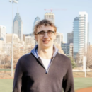 A person with glasses smiling in front of a cityscape featuring modern skyscrapers and a sports field.