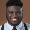 Smiling headshot of a person wearing a light blue shirt, black tie, and suspenders against a brown background.