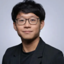 Headshot of a person with short dark hair and glasses, wearing a black jacket and a black shirt, against a plain background.