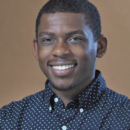 Smiling person in a headshot wearing a dark, polka-dotted shirt, with a neutral brown background.