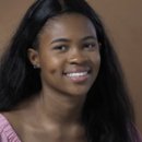 Headshot of a person smiling with long dark hair, wearing a pink top, against a brown background.