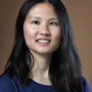 Headshot of a person with long, dark hair wearing a blue shirt, smiling at the camera against a neutral background.