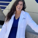 A person smiling while wearing a white lab coat, likely indicating a professional in a medical or scientific field. They're standing outdoors with a blue and white structure in the background.