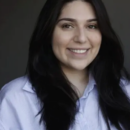 A person with long dark hair smiling, wearing a light-colored collared shirt, set against a dark, neutral background. Headshot photo.