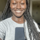 A person with long braided hair smiling while taking a selfie. They are wearing a gray shirt and standing in bright sunlight indoors.