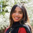 A person smiling outdoors with blooming trees in the background, wearing a black scarf and red clothing, layered gold necklaces visible.