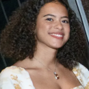 A person with curly hair smiling, wearing a white and gold patterned outfit and a necklace.