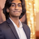 A headshot of a person with long hair wearing a suit jacket and shirt, smiling slightly, standing against a blurred, warmly-lit background.