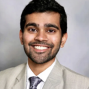 A person wearing a light-colored suit and tie, smiling in a professional headshot with a neutral background.