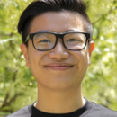 A headshot of a person wearing glasses and a black top, smiling outdoors with greenery in the background.