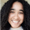 Headshot of a person with curly hair, smiling and looking at the camera, wearing a light-colored top.