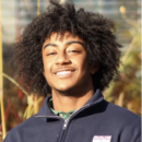 A person with curly hair smiling, wearing a dark jacket with a logo, standing outdoors in a sunlit environment with plants in the background.