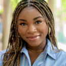 A headshot of a person with braided hair, smiling, wearing a blue striped shirt. The background is soft-focus greenery.