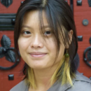 Headshot of a person with medium-length hair and highlights, wearing a gray shirt and green earrings. The background features a red door with ornate black detailing.