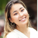 Headshot of a smiling person with shoulder-length hair wearing a white top and hoop earrings, set against a blurred outdoor background.