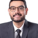 Headshot of a person wearing glasses and a suit with a tie, smiling against a plain white background.