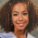 A headshot of a person with curly hair and decorative hairpins, smiling, wearing earrings and a gown indicating a formal or graduation setting.