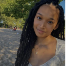 A person with long hair is smiling softly while on a sandy beach. They are wearing a light-colored top and a necklace with a pendant. Trees are visible in the background.