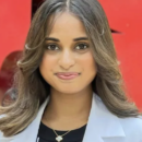 A headshot of a person with shoulder-length hair, wearing a white coat and a necklace with a small pendant. In the background, there is a large red structure.