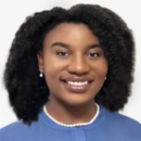 Headshot of a person with curly hair, smiling, wearing a blue top and pearl earrings, set against a plain background.