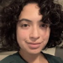 Headshot of a person with curly dark hair, wearing a green top, smiling slightly with a neutral background.