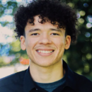 A person with curly hair smiling, wearing a dark shirt, standing in front of a blurred background with green foliage. Headshot.