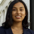 A person with shoulder-length hair wearing a dark blazer and necklace, standing in front of a building with white columns.