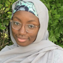 A person wearing glasses and a light gray hijab, standing in front of green foliage. They have a patterned headband visible under the hijab.