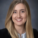 This is a professional headshot of a person with long, blonde hair, smiling, wearing a dark blazer and a light-colored top. The background is a neutral gray.