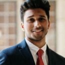 A headshot of a person wearing a suit and a red tie, smiling in a well-lit, formal setting.