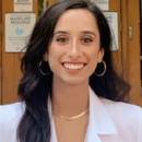 Headshot of a person with long dark hair, smiling and wearing a white coat with a logo on it. Visible signs in the background indicate masks are required.